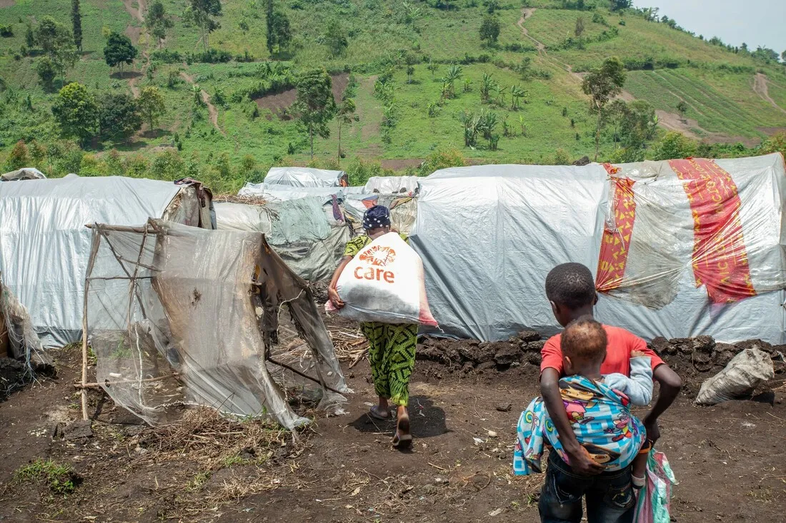 A woman walks toward a makeshift shelter made of plastic sheets, carrying a sack with the CARE logo. A young boy follows her, carrying a smaller child tied to his back; they are likely her children.