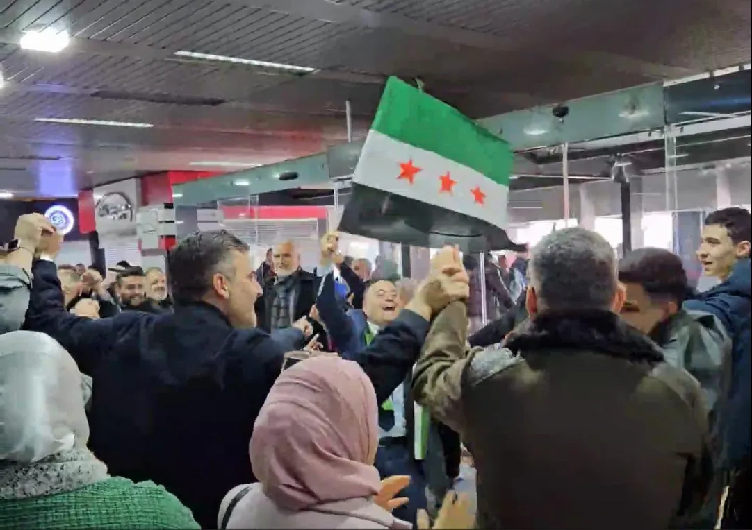 People dance and sing in a circle at Damascus airport, one person at the center waving a Syrian flag.
