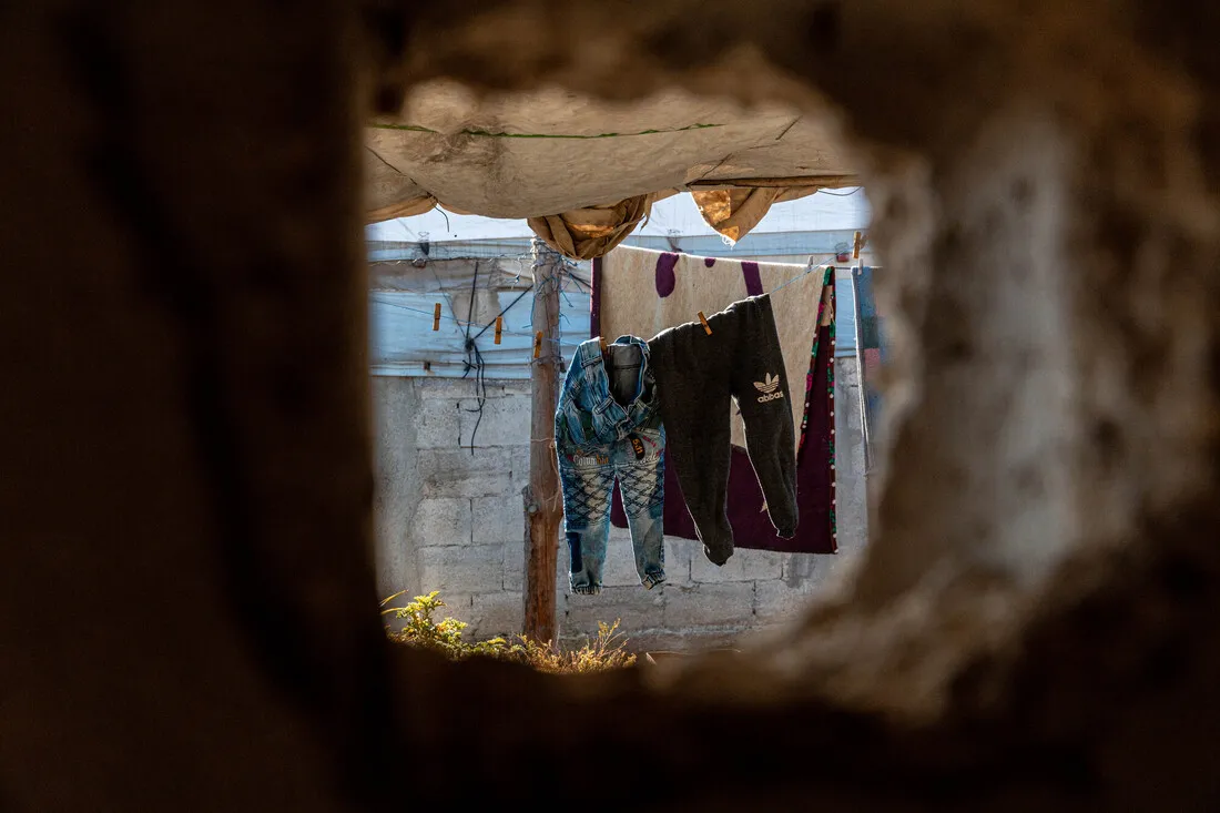 A large, round hole in a wall, likely caused by shelling, frames a view of a courtyard with clothes drying in the sun.