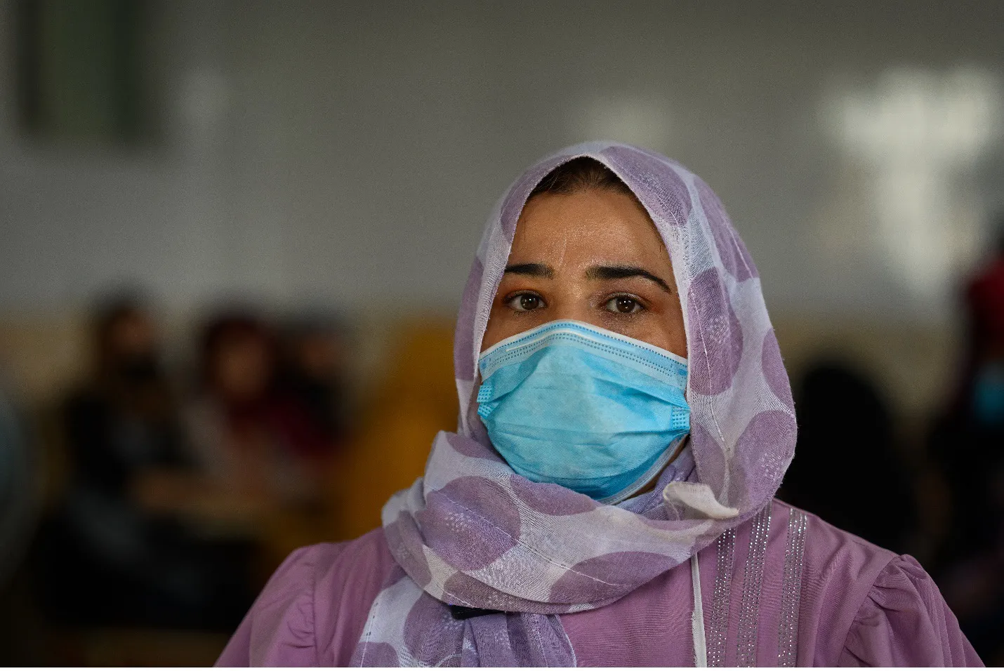 Medium portrait of a woman in mask, head scarf, and purple clothing, looking at the camera.