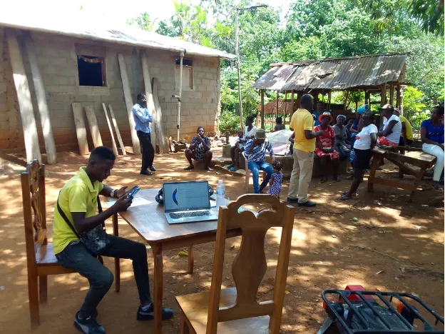 Outdoor photo of a man working on a smartphone and laptop computer at a table, with others seated and standing in the background.