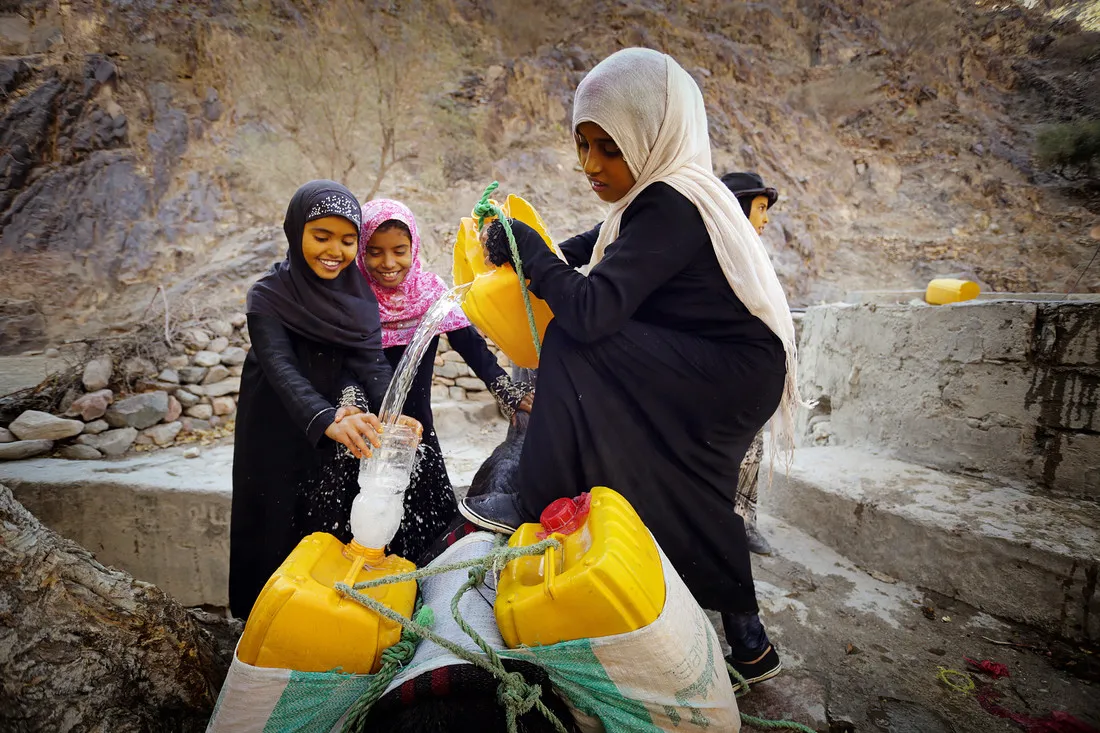 A girl pours water from a bucket attached to a well into water jugs.